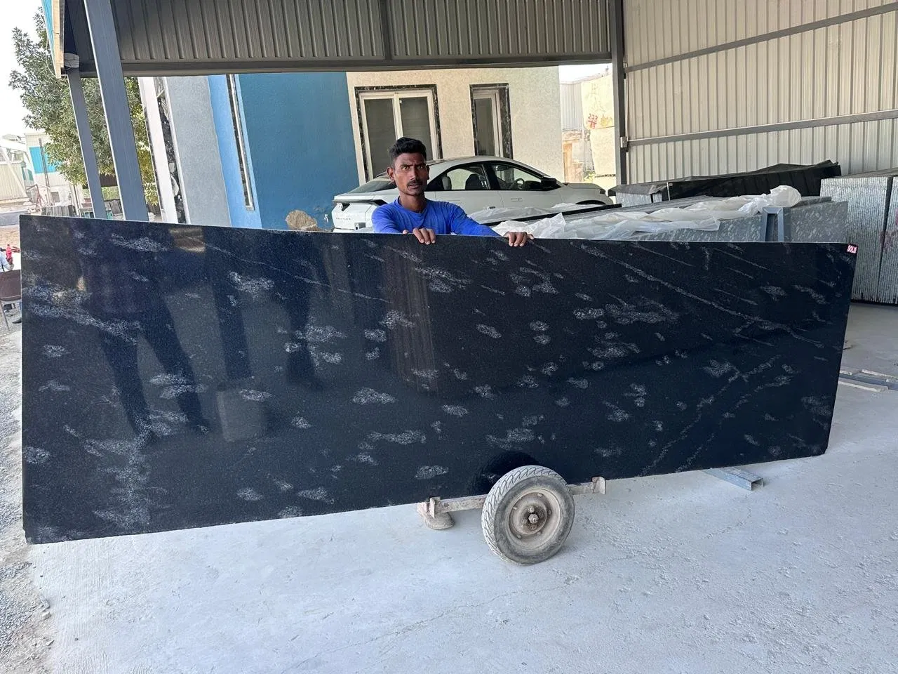 Nature’s Art: Incorporating Fish Black Granite in Artistic Architectural Installations 2 A worker handles a large, polished black granite slab with white and grey specks at a covered processing facility. The slab is on a wheeled dolly, highlighting precision cut and bulk supply capability.